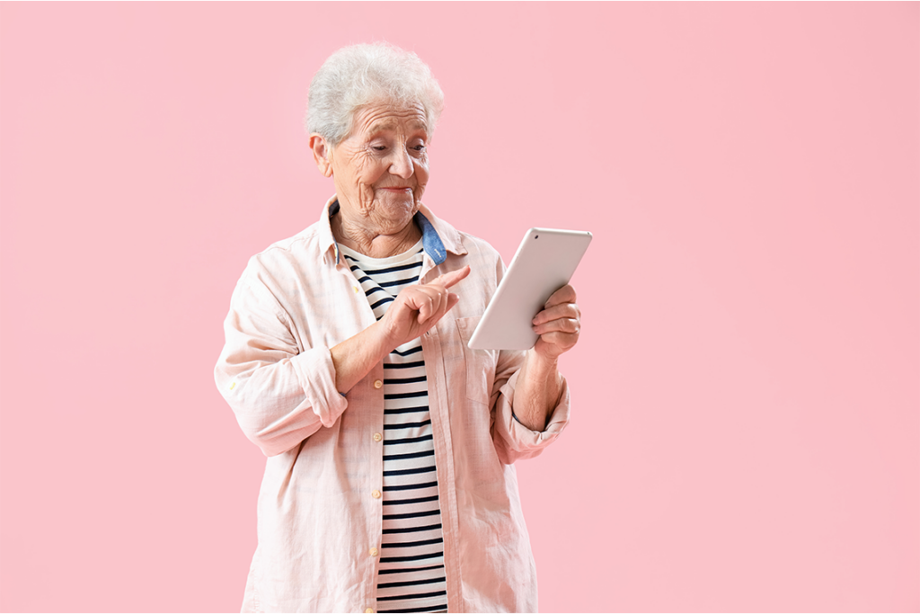 An older woman with white hair stands in front of a pink background, scrolling on a tablet and smiling.