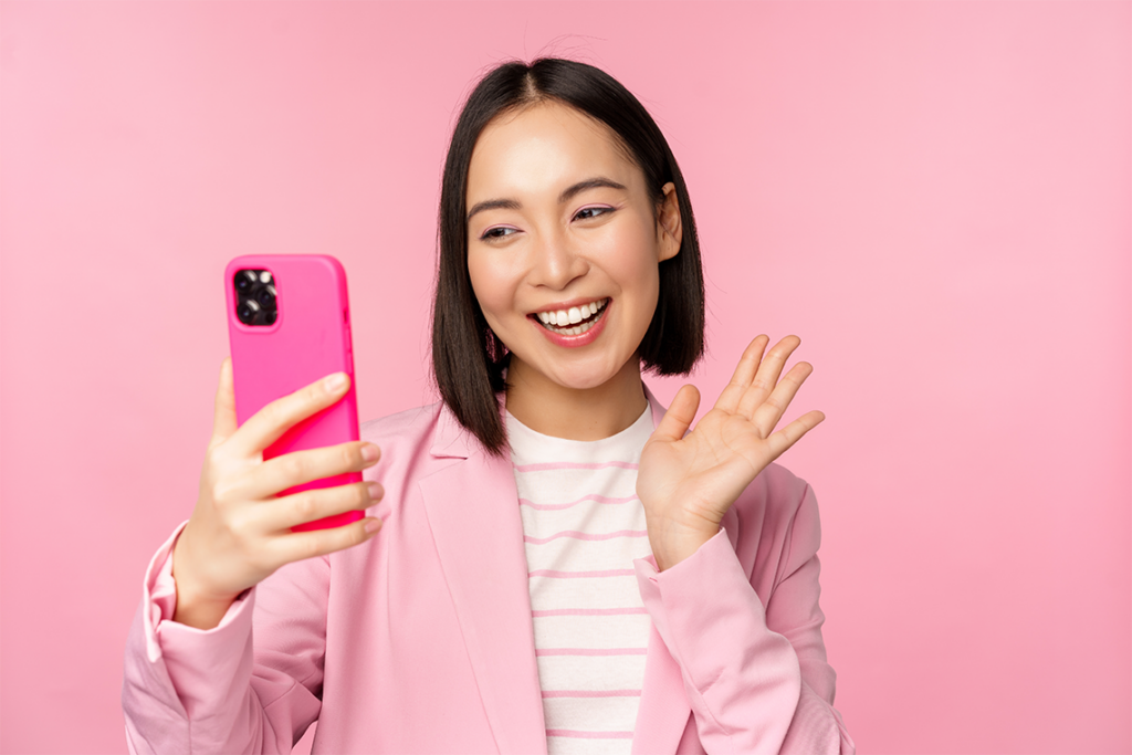 A woman with a dark bob hairstyle, wearing a striped pink and white shirt and a pink blouse, smiles and waves at her smartphone screen. The phone has a hot pink case.