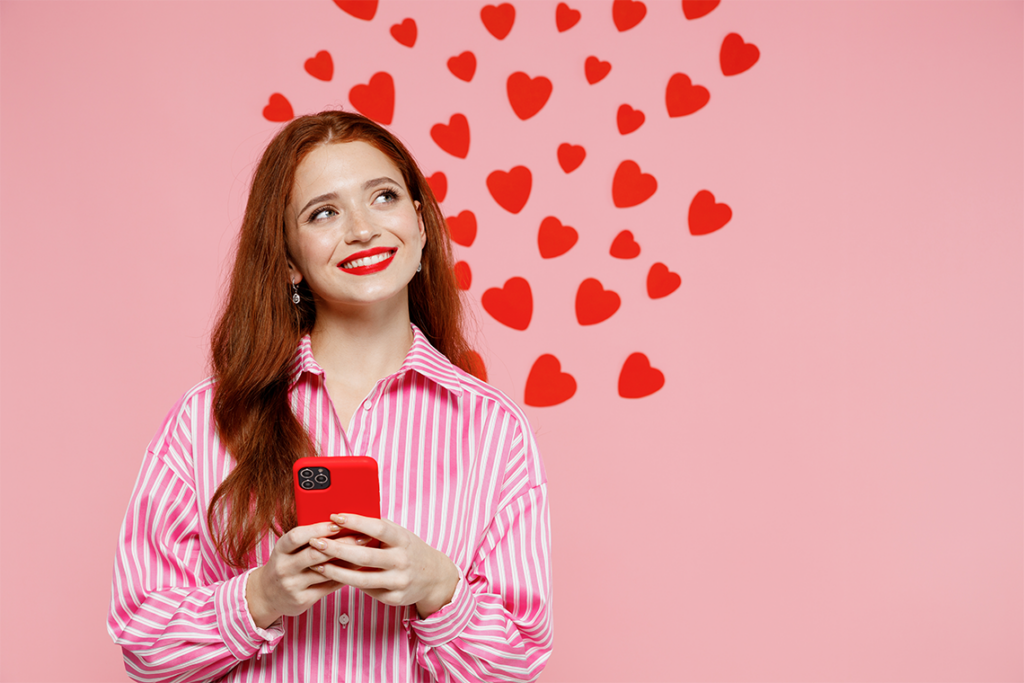 A smiling woman with long red hair and wearing a pink striped shirt holds a smartphone and looks up in thought. Pink background with red hearts.