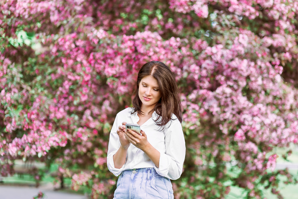 A woman in a white blouse and jeans stands in front of a shrub full of pink flowers, looking down at her phone.