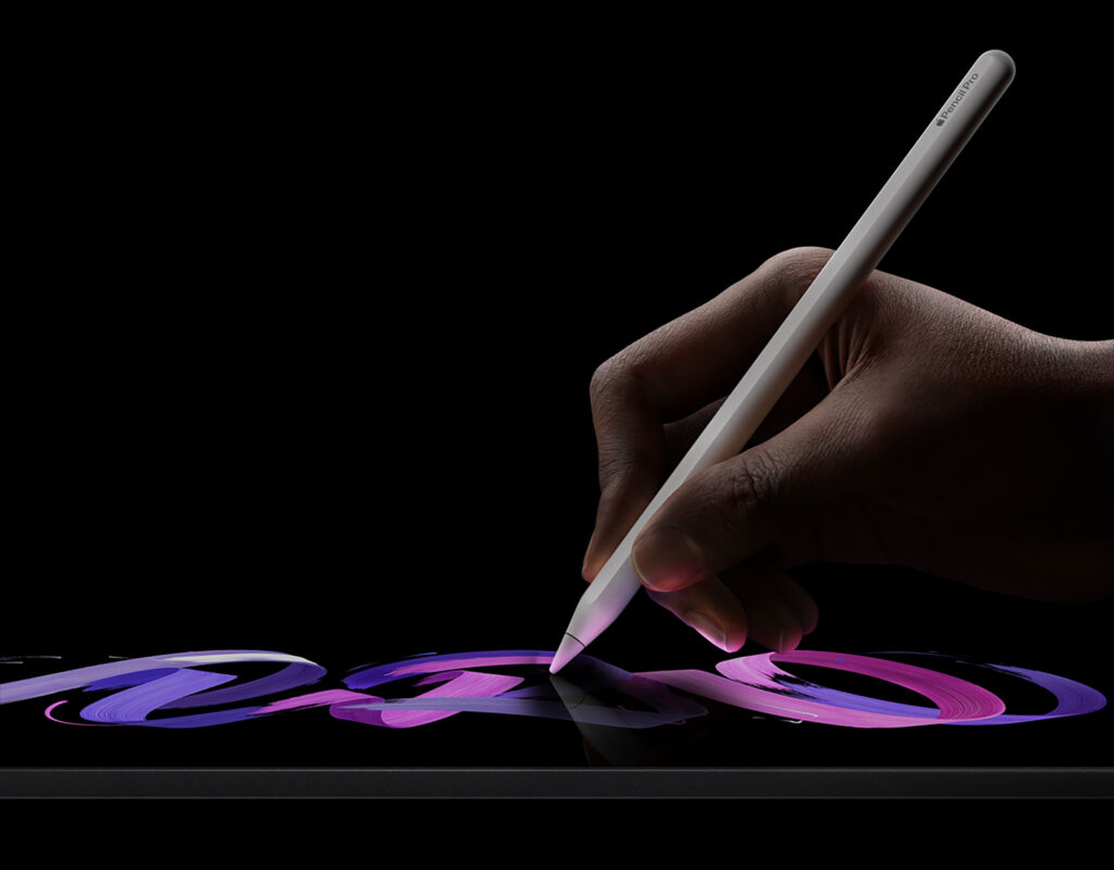 A hand against a black background holds a stylus over a lit 2024 iPad Pro tablet.