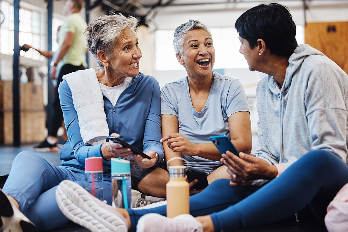Three elderly women in workout clothing sit on a floor together, smiling and talking while holding their smartphones.