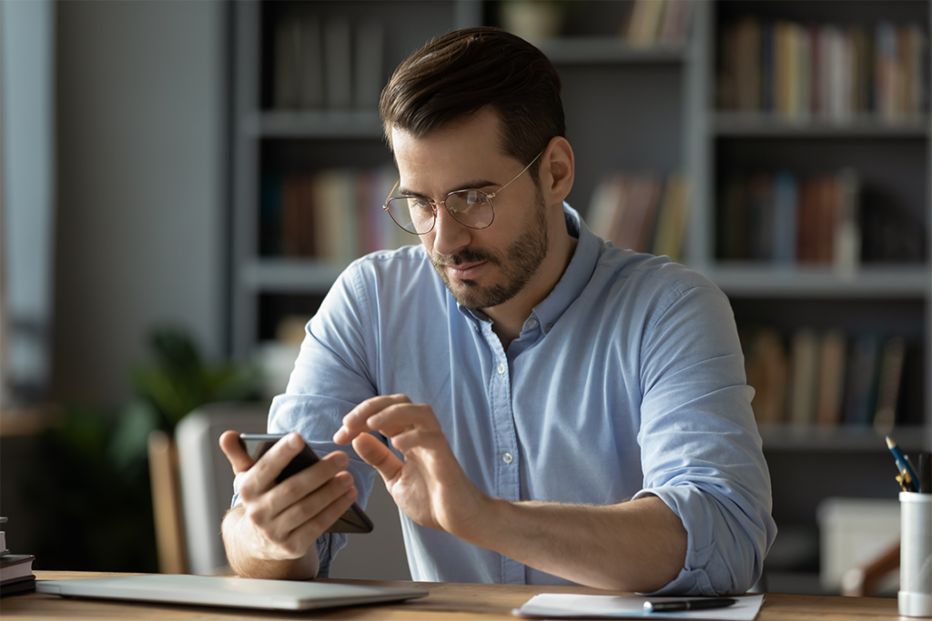 A man in a light blue dress shirt sits at a desk, looking down at this smartphone. He has short brown hair and a beard, and is wearing glasses. A bookshelf full of books, a plant, and chairs are visible in the background.