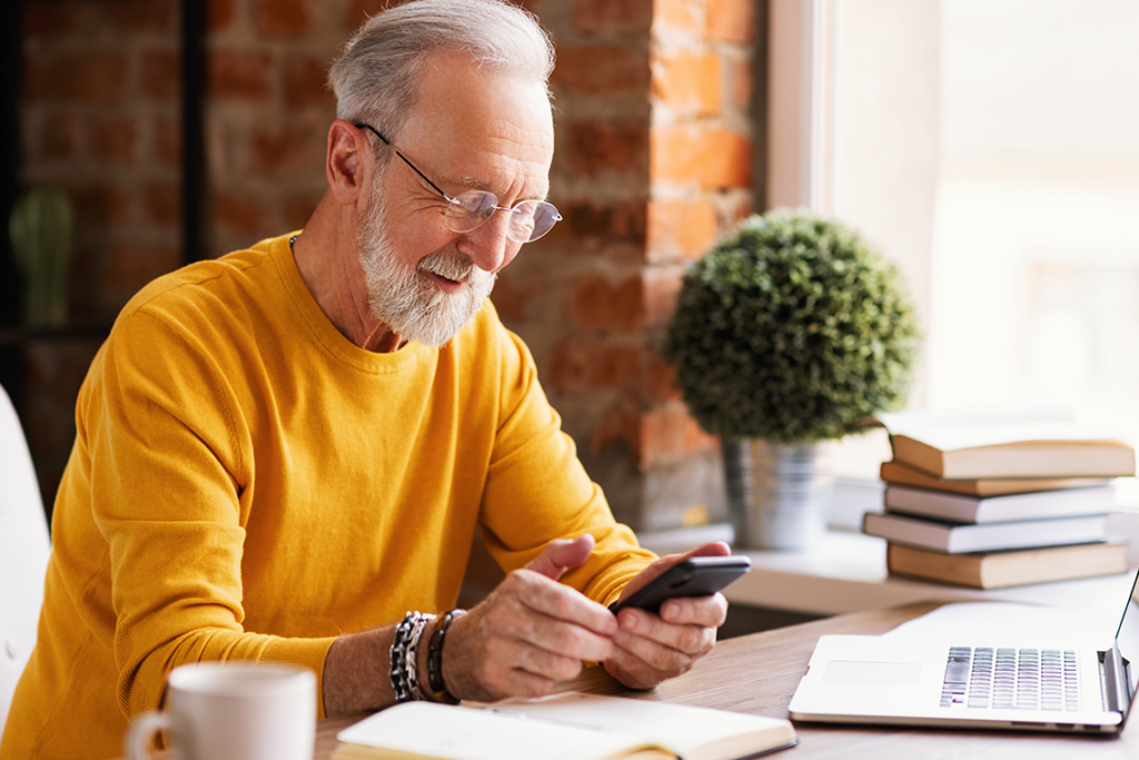 An elderly man with silver hair and beard, wearing glasses and a yellow sweater, sits at a desk near a window. He's smiling and looking down at his smartphone. The desk is covered in books, along with a coffee mug, laptop, and potted plant. 