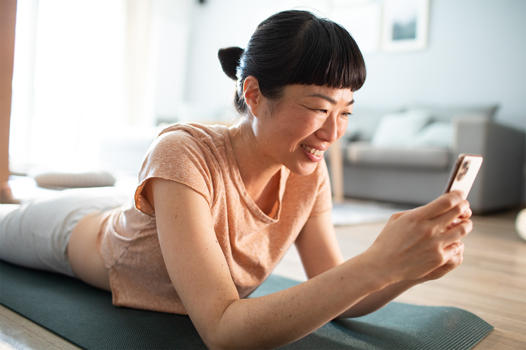 A woman with black hair tied back in a bun lays on her stomach on top of a yoga mat in her living room. She's propped up on her elbows, holding a smartphone and smiling down at it.