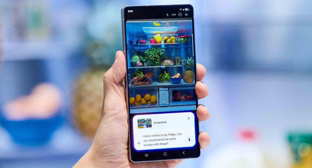 A Samsung Galaxy S25 held in hand in front of an open refrigerator. The display shows the contents of the fridge with a Google Gemini text screen request for recipes.