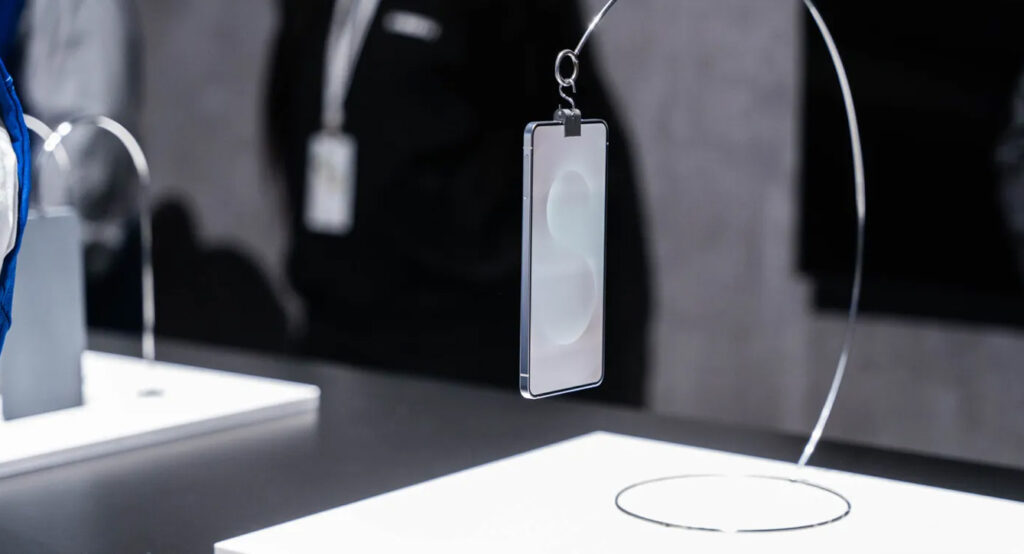 Samsung Galaxy S25 Edge, dangling from a mount above a white panel on a gray tabletop.