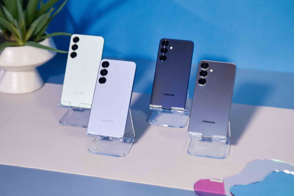 A group of Samsung Galaxy S25+ smartphones, on short clear pedestals. The pedestals sit on a tabletop with a potted plant and blue wall in the background.