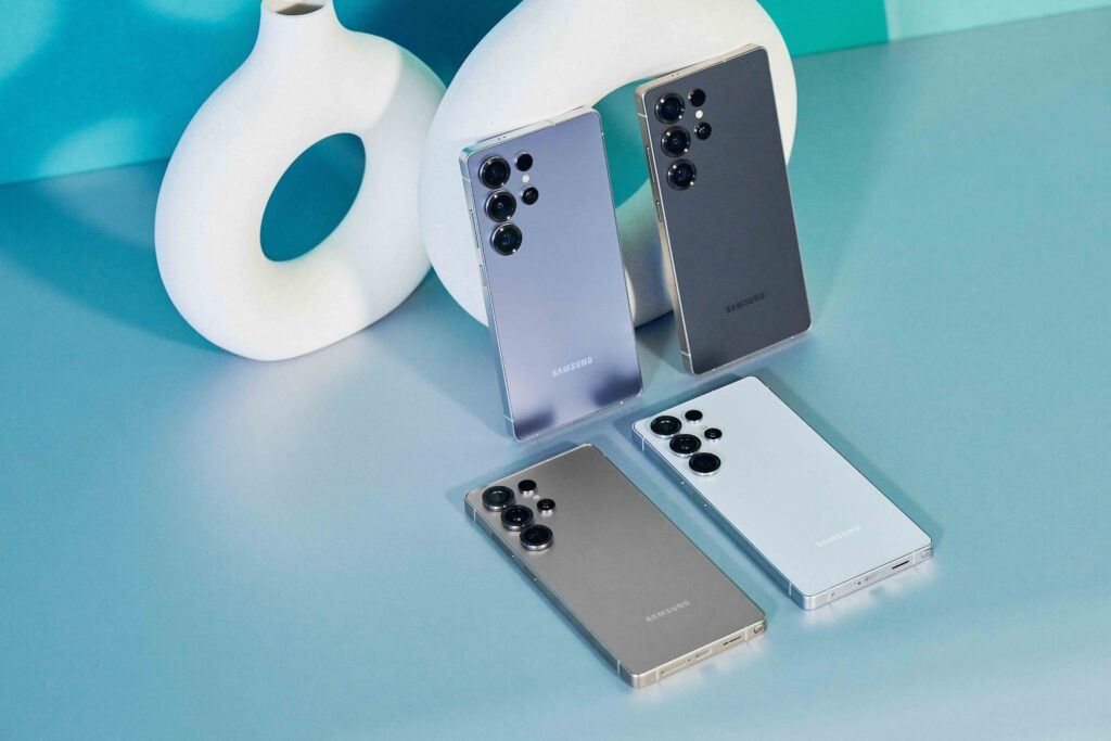 A group of Samsung Galaxy S25 Ultra smartphones, two standing and two laid flat on a blue-green surface. 