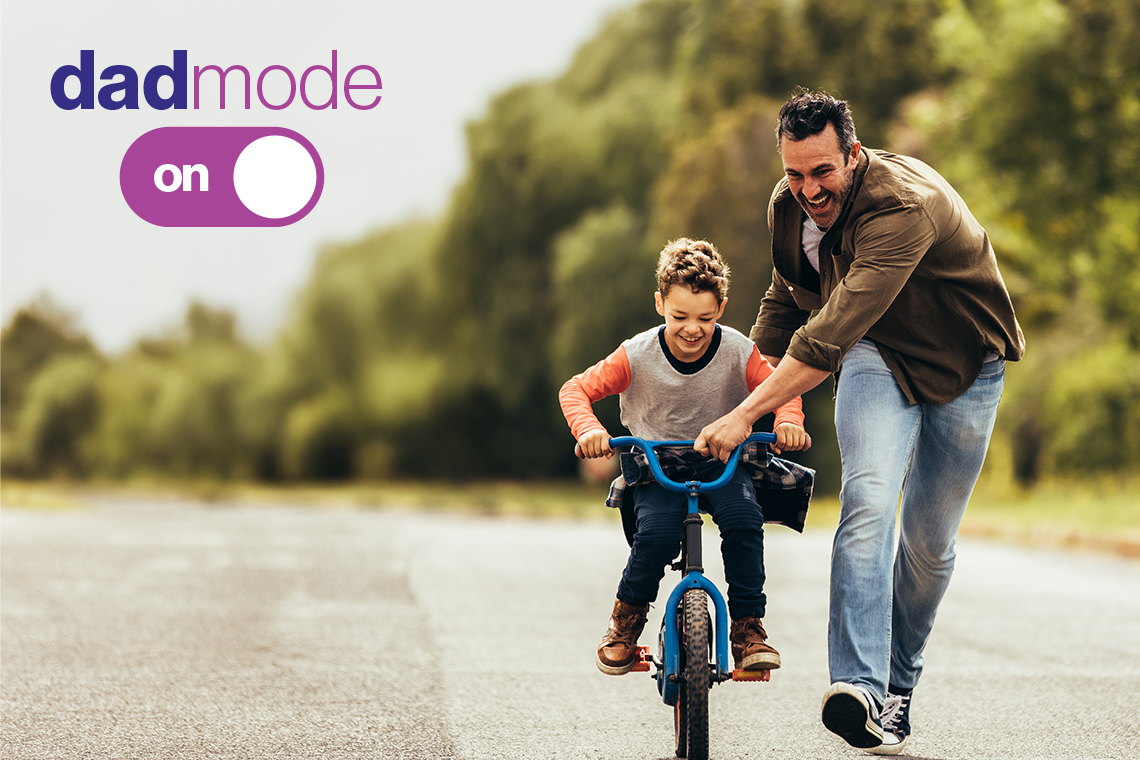 A father in a brown jacket and jeans helps his son, wearing a gray and orange teeshirt and dark jeans, ride a bike. In the top left corner is a Father's Day theme logo, reading "Dad Mode" above a toggle switch that reads "on."